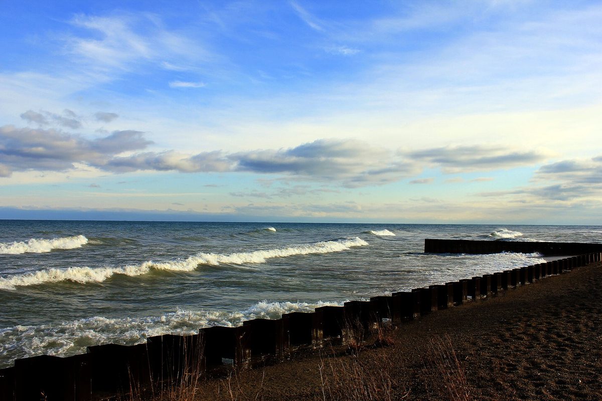 Waves breaking along a weathered wooden groin on a sandy Lake Michigan shoreline in Illinois under a partly cloudy sky.