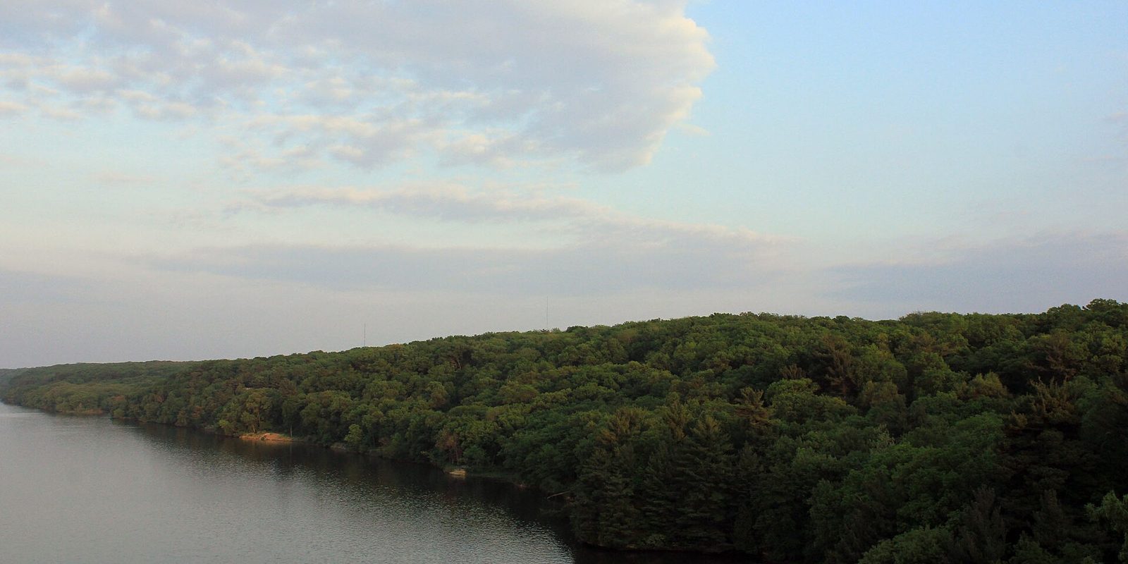 A broad calm river curving past a long wooded bluff under a pale, cloud-streaked sky.