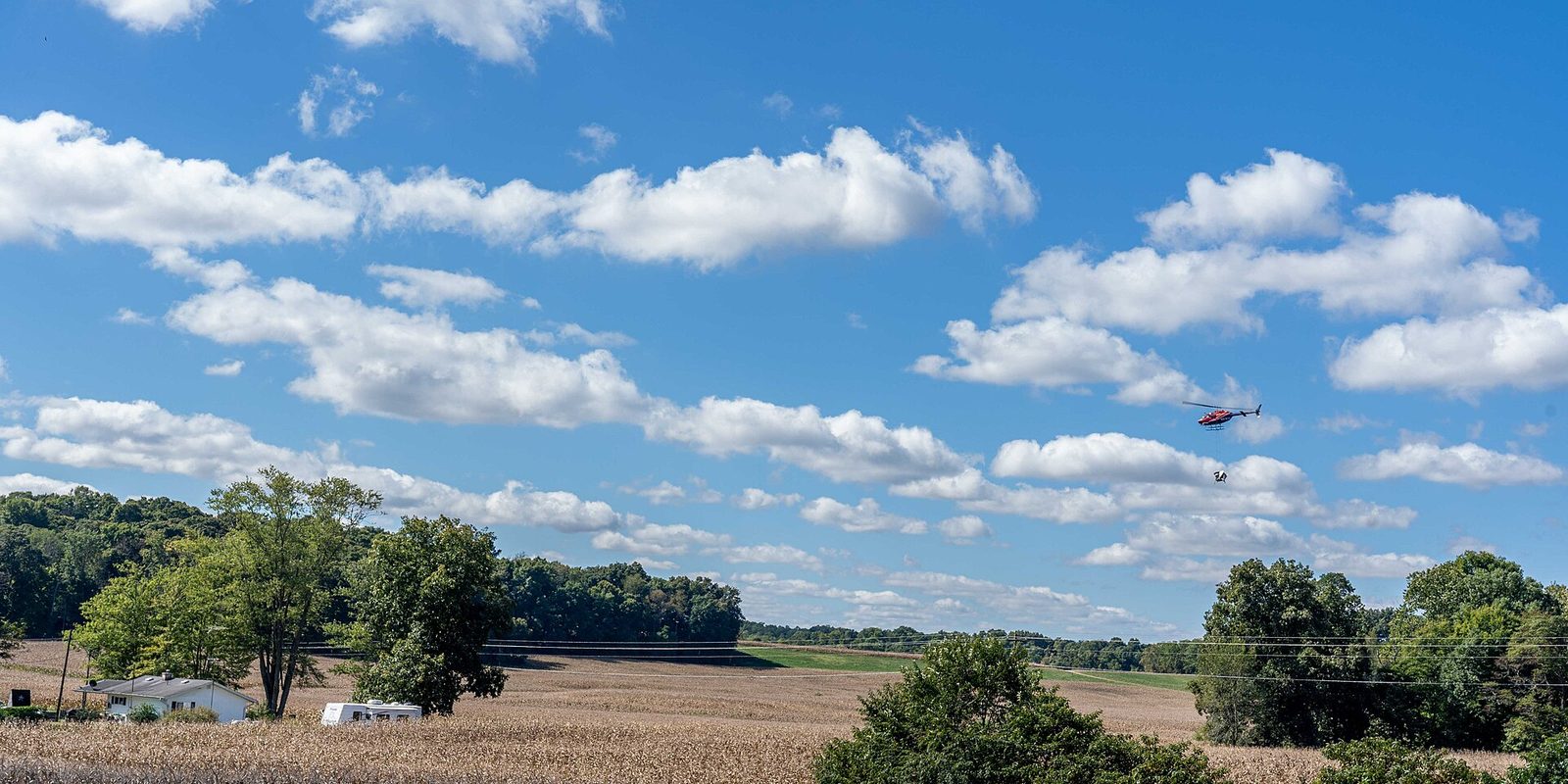A helicopter hovers over harvested farm fields, trees and a small house under drifting clouds