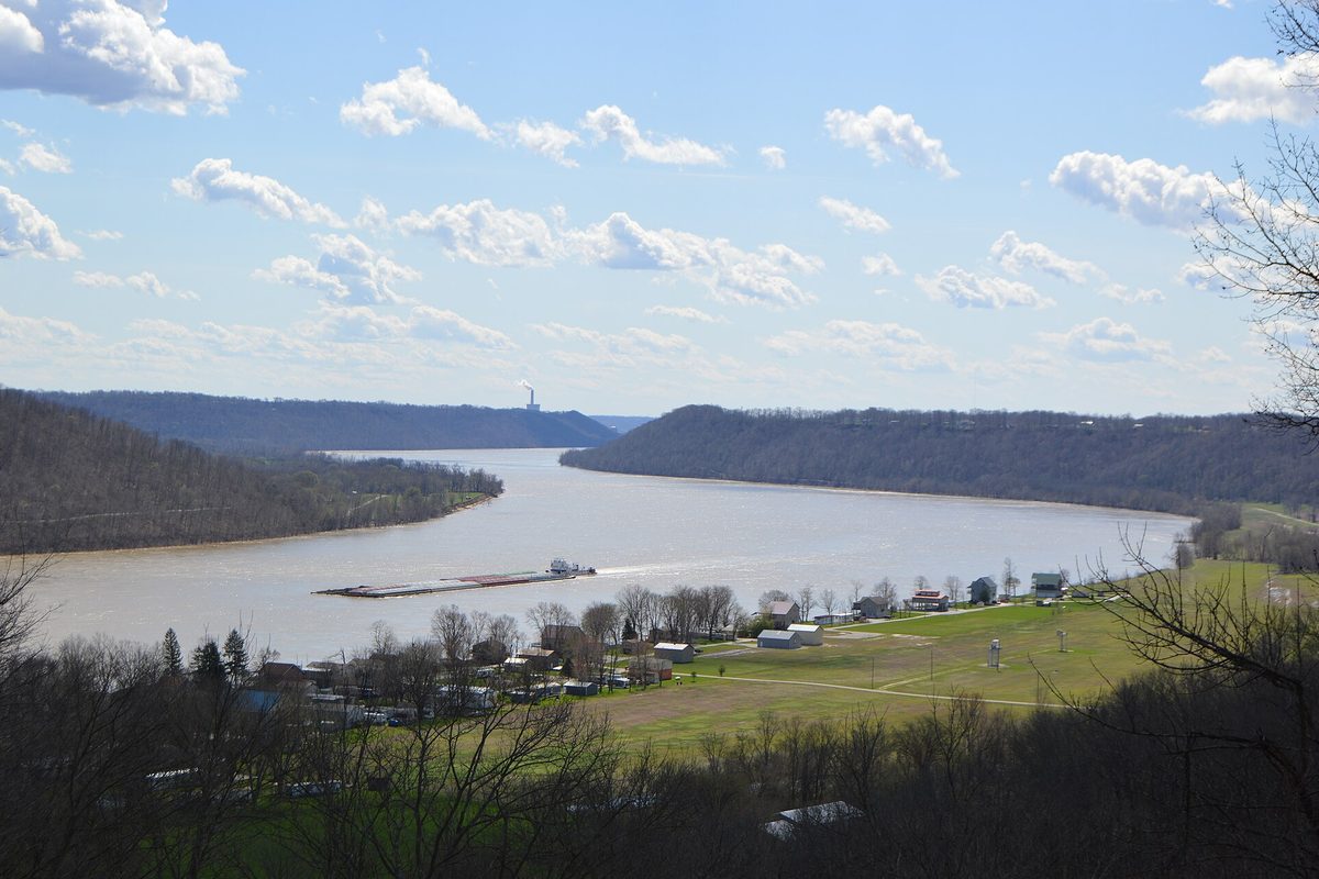 Towboat pushing a loaded barge around a bend of the Ohio River in Kentucky, past wooded bluffs and a small riverside town.