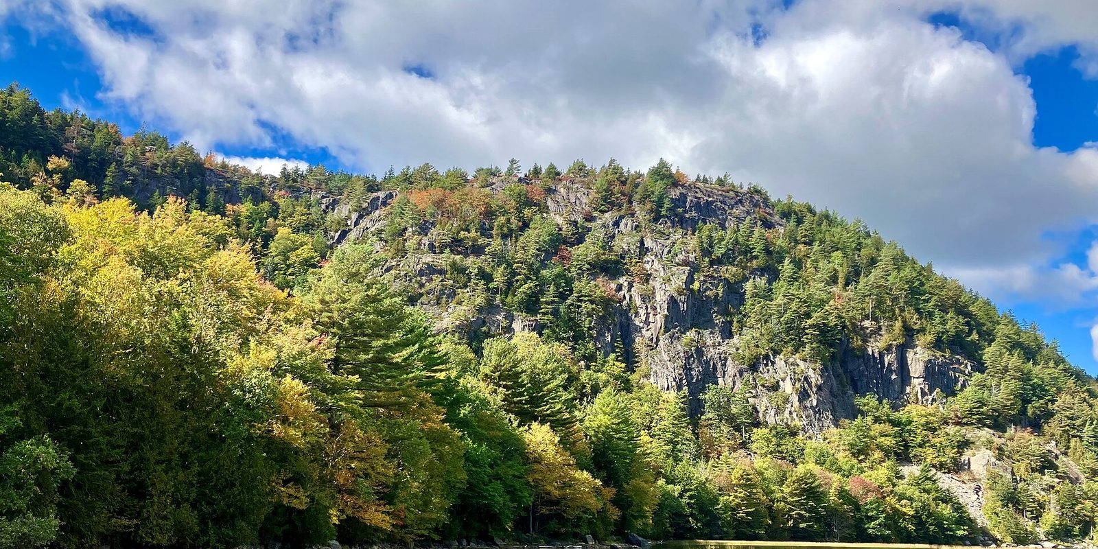 Rocky wooded hillside with early autumn colour rising above trees under a bright cloudy sky