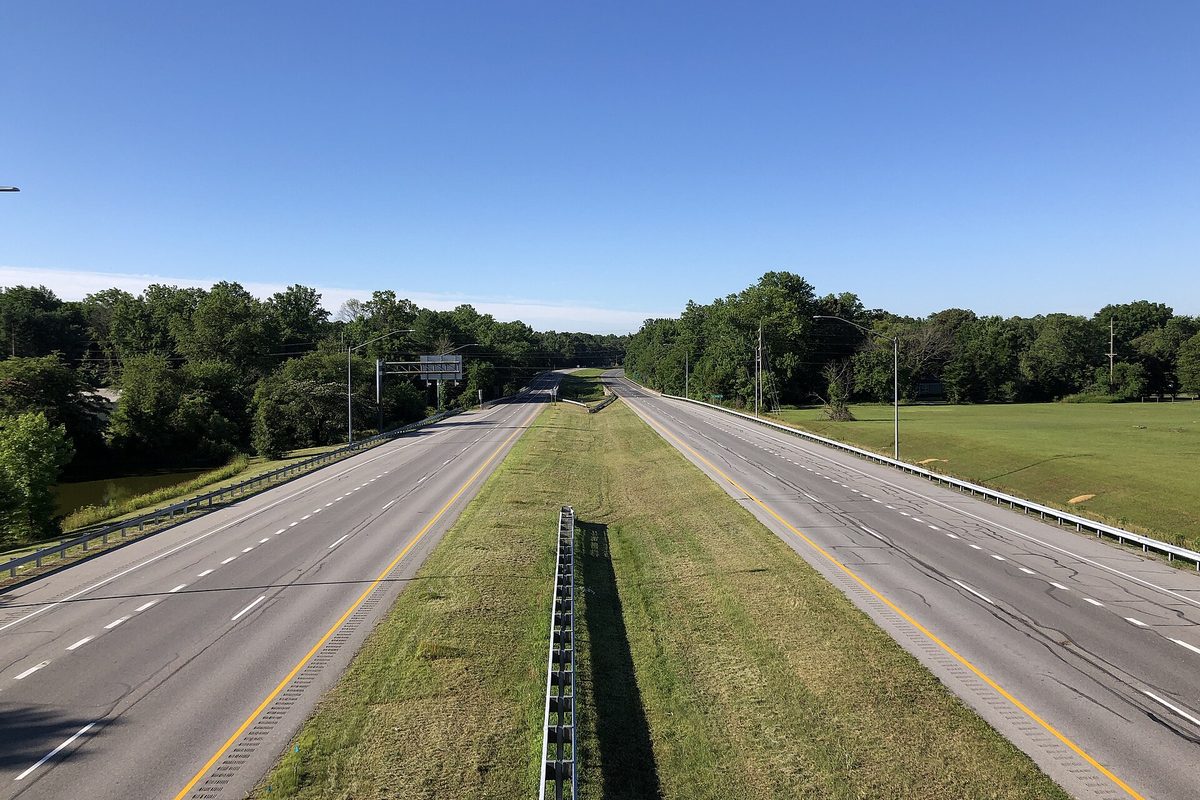 Empty divided highway in Maryland seen from an overpass, both roadways running toward woods past a mowed grass median under a clear blue sky.