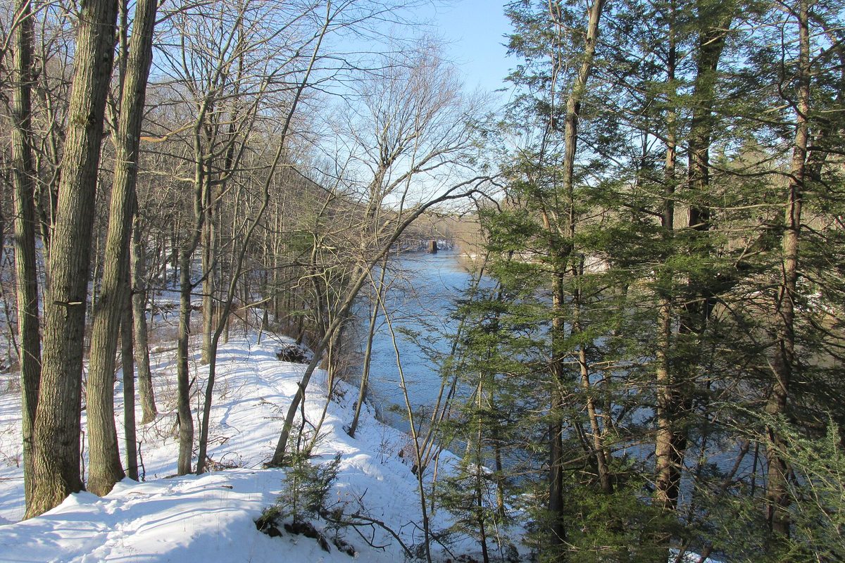 Snow-covered wooded bluff in Massachusetts dropping to a blue river, bare hardwoods and hemlocks framing an old stone pier downstream.