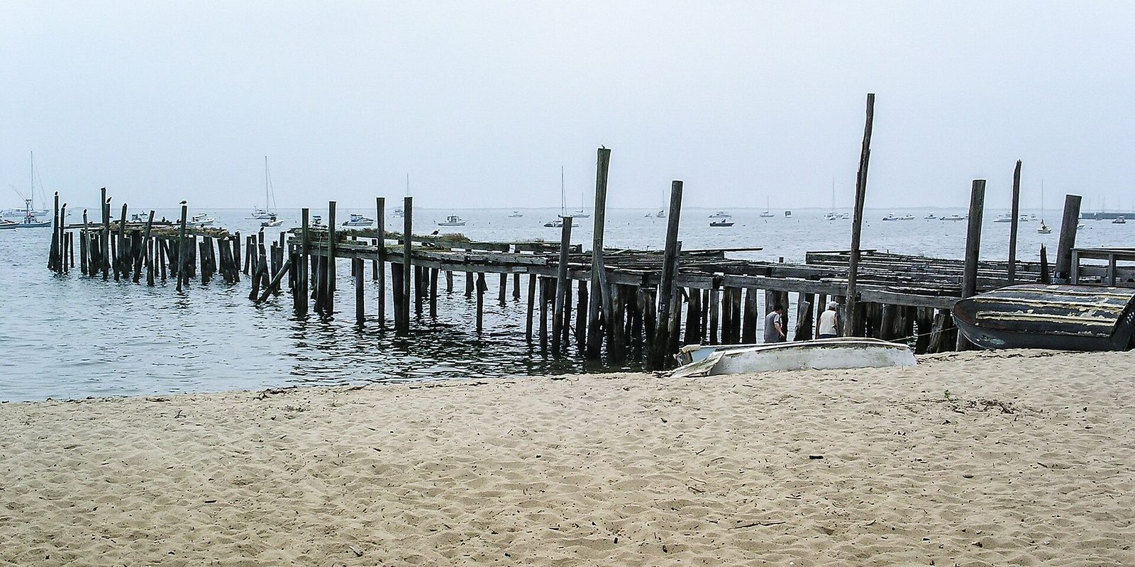 Two people stand beside a collapsing wooden pier on a sandy beach, sailboats moored in hazy water beyond.