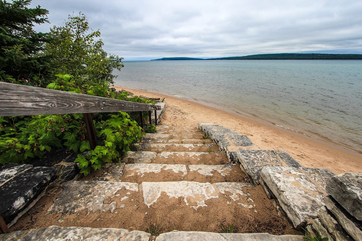 Weathered stone steps and a wooden handrail dropping to a sandy Michigan lakeshore, with a forested point across the water under an overcast sky.