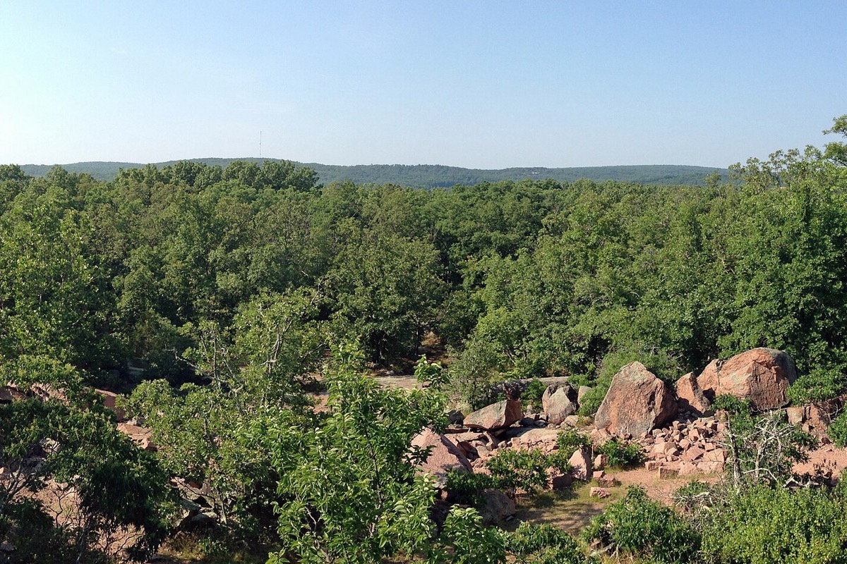 Rounded pink granite boulders lying in dense green Missouri woodland, with more of them scattered over the slope and a long wooded ridge running away behind.