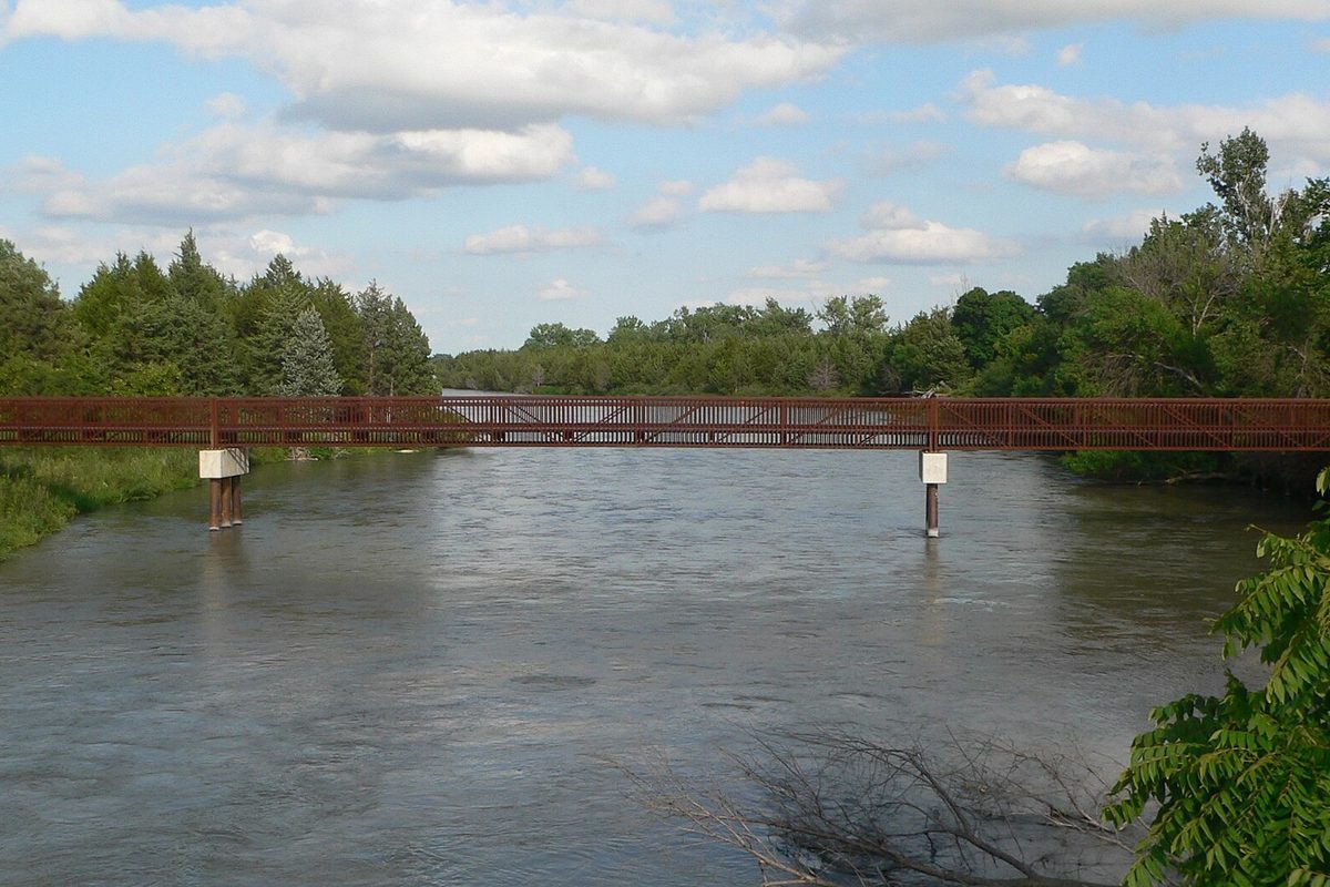 Nebraska: a rust-red steel truss footbridge on concrete piers crosses a wide river lined with pines, under puffy cumulus clouds.