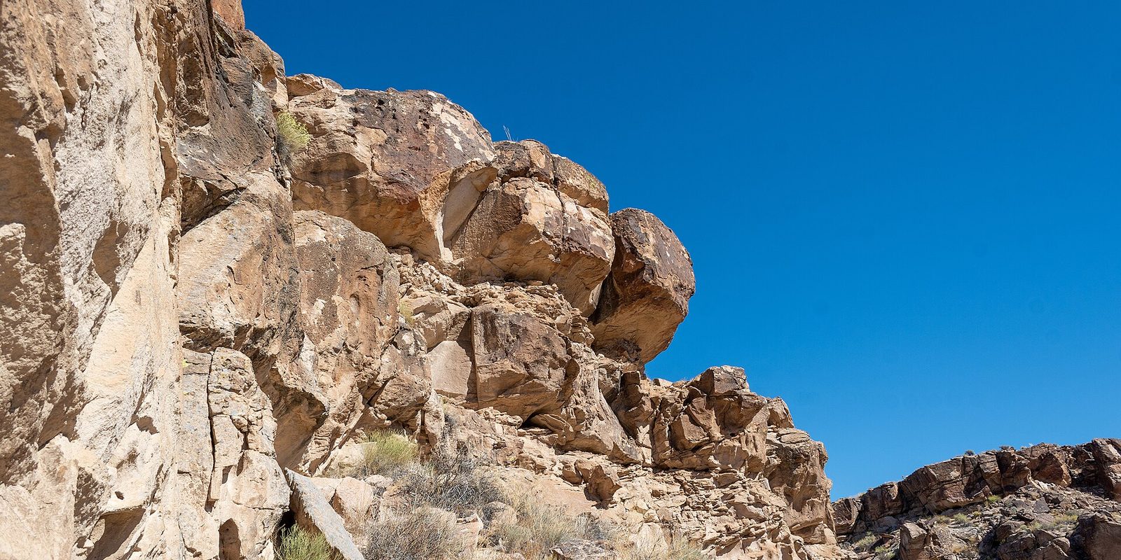 Sunlit tan rock cliffs and boulders topped with dry scrub under a deep blue sky