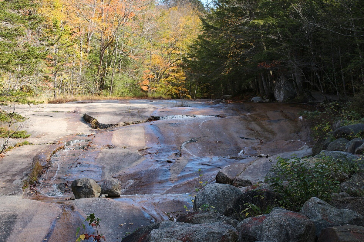 A New Hampshire brook running in shallow sheets over a broad slab of smooth pink rock, boulders along the near edge and hardwoods turning yellow on the bank behind.