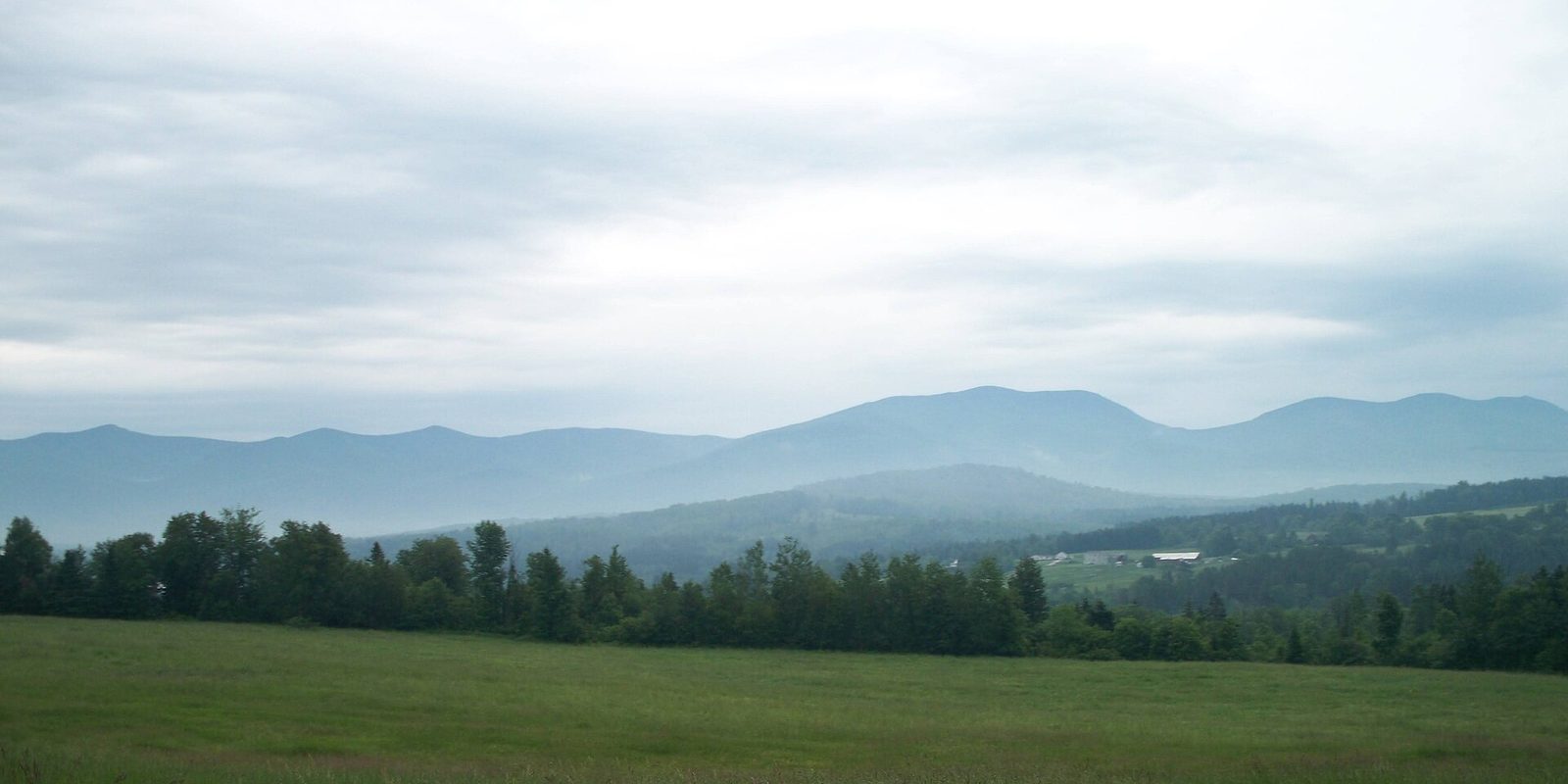 A mown green field and tree line below layered blue mountain ridges under hazy overcast sky