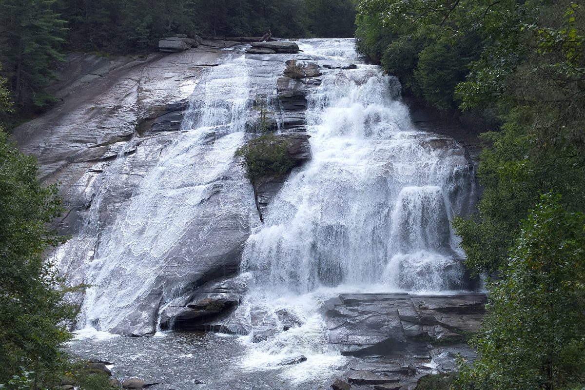 North Carolina waterfall spilling in two white channels down a broad tilted granite face, hemmed in by dense green forest.