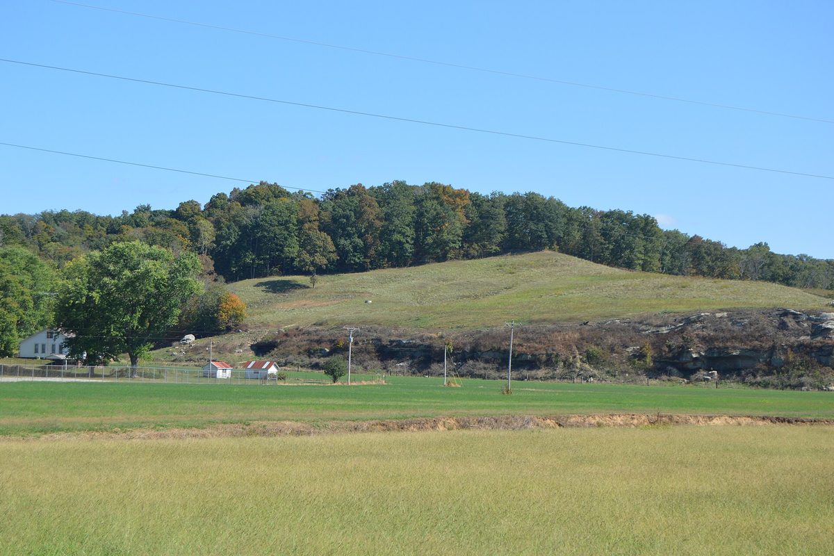Ohio farm fields below a wooded ridge, with a white farmhouse, a red-roofed shed and an exposed sandstone ledge under blue sky.