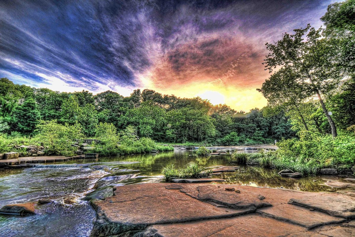 Oklahoma creek stepping over low rock ledges into small falls, flat stone slabs in front and green woods under a fiery sunset.