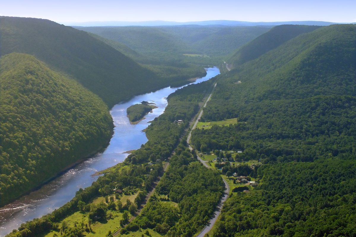 Aerial over a Pennsylvania river valley, where a wide blue river bends between steep forested ridges past a small village and a two-lane highway.