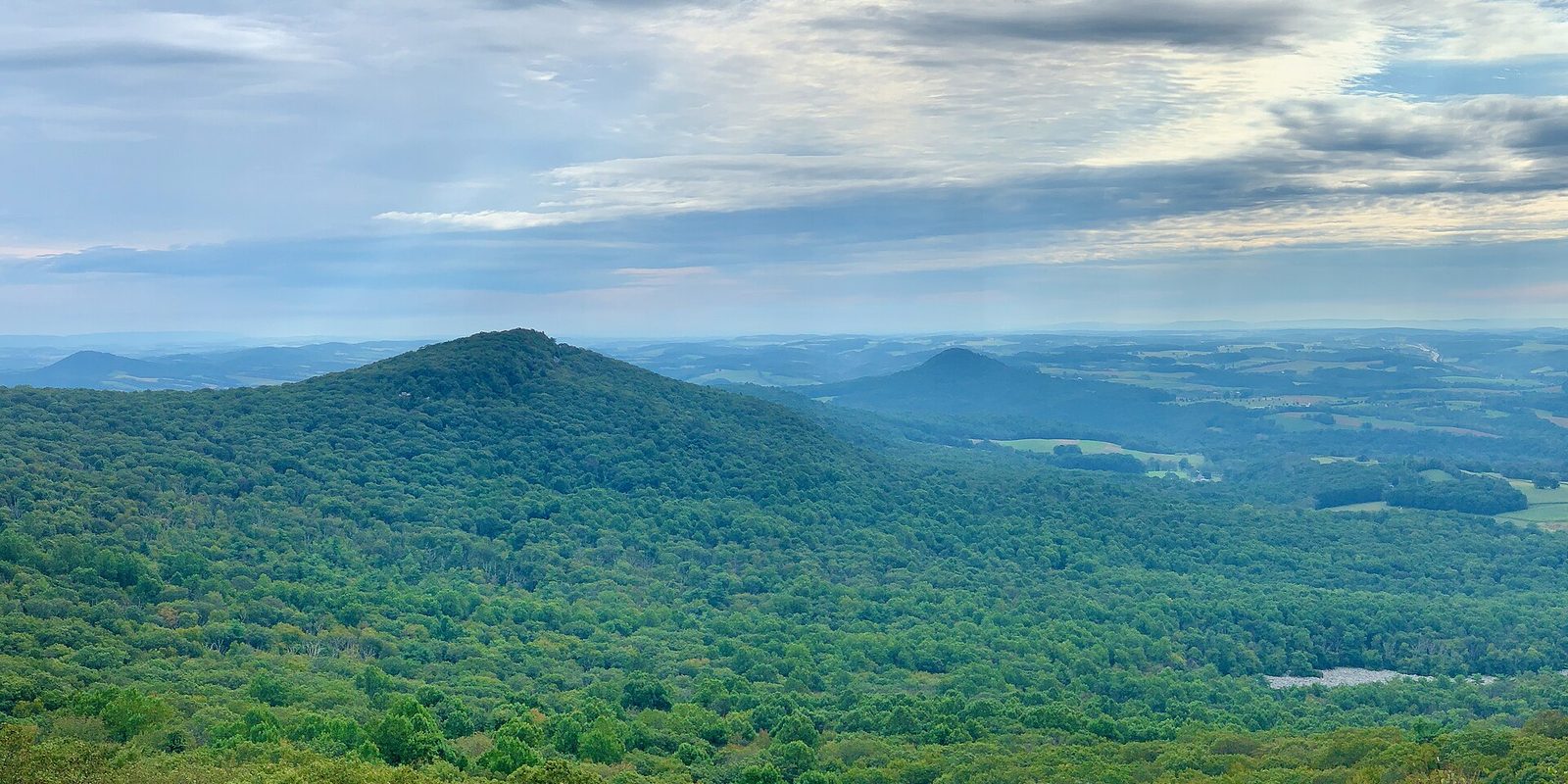 Rounded forested hills above a wide valley of farm fields, under soft banded clouds