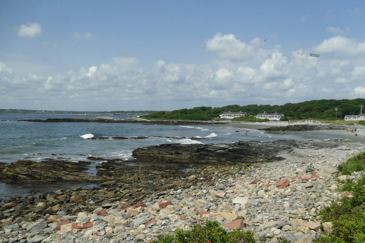 A Rhode Island shore of flat rock shelves and rounded cobbles with small surf breaking over them, a low green headland and a few white houses along the point beyond.