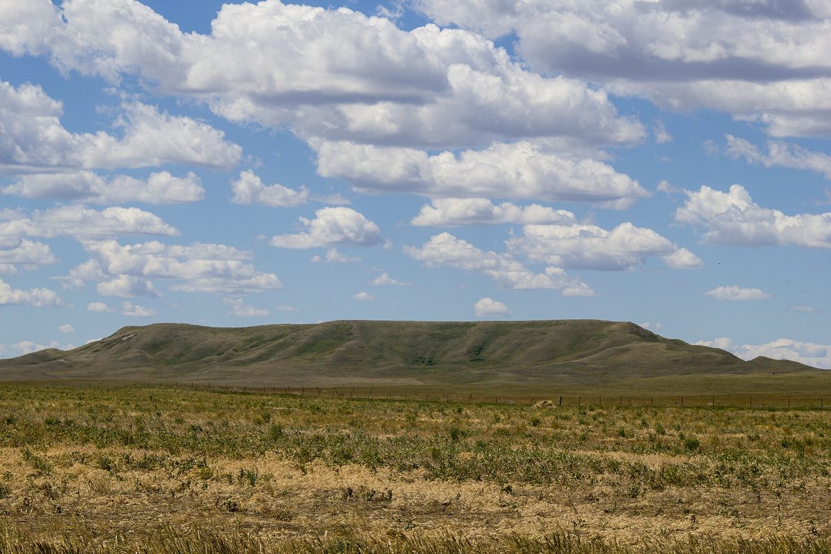 South Dakota grassland, where a long low grass-covered butte rises beyond a barbed-wire fence line under scattered cumulus clouds.
