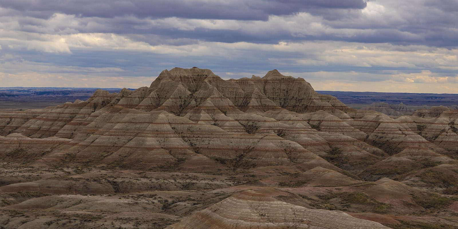 Banded, eroded badlands ridges under a heavy grey cloud bank, flat plains stretching behind