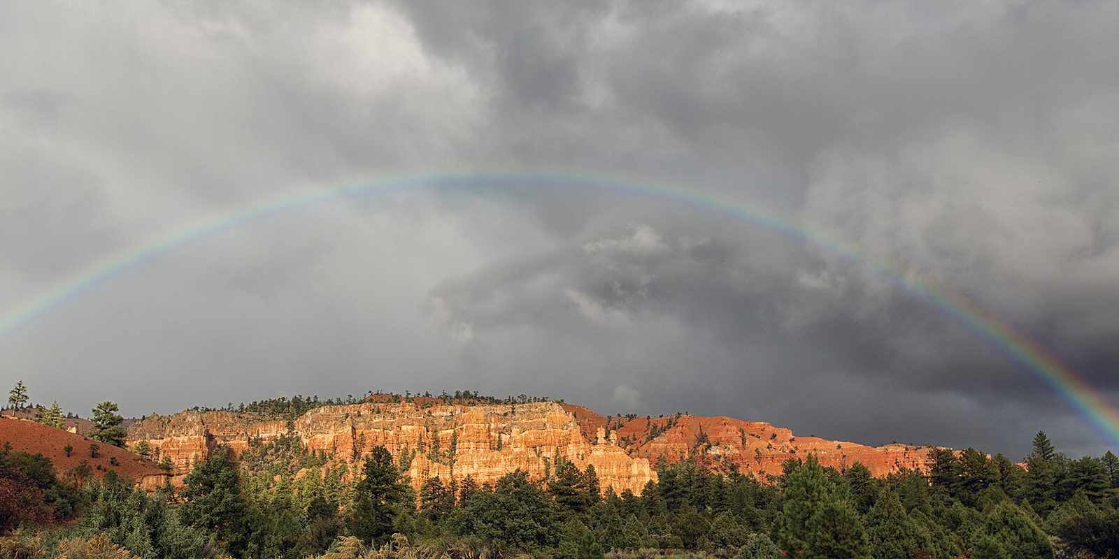 A faint rainbow arcs over red rock cliffs and pine trees under heavy grey storm clouds.
