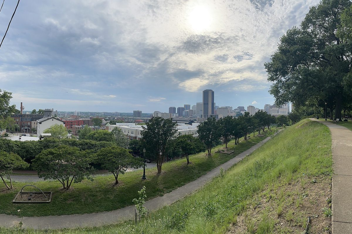 Richmond's downtown towers in Virginia rising past rooftops beyond a grassy hilltop park, a paved path curving down the slope
