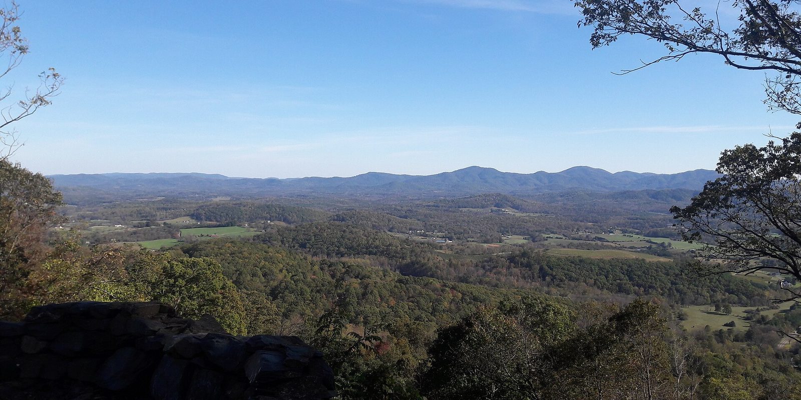 Rocky ledge and leafy branches framing wooded valley farmland below a hazy blue mountain ridge