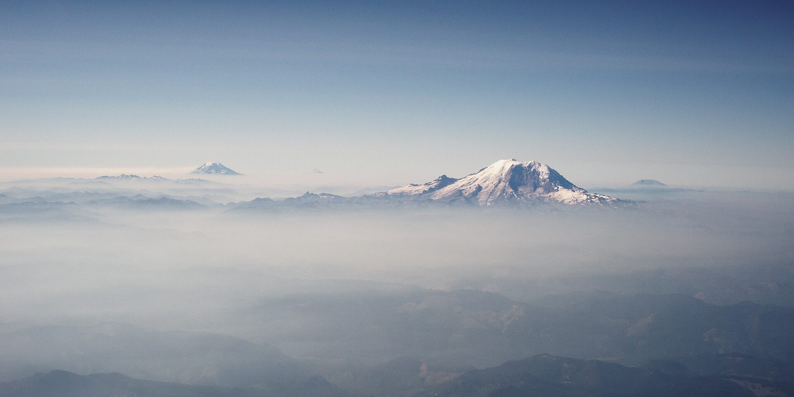 A snow-capped volcanic peak rising above layers of haze and low ridges under pale sky.