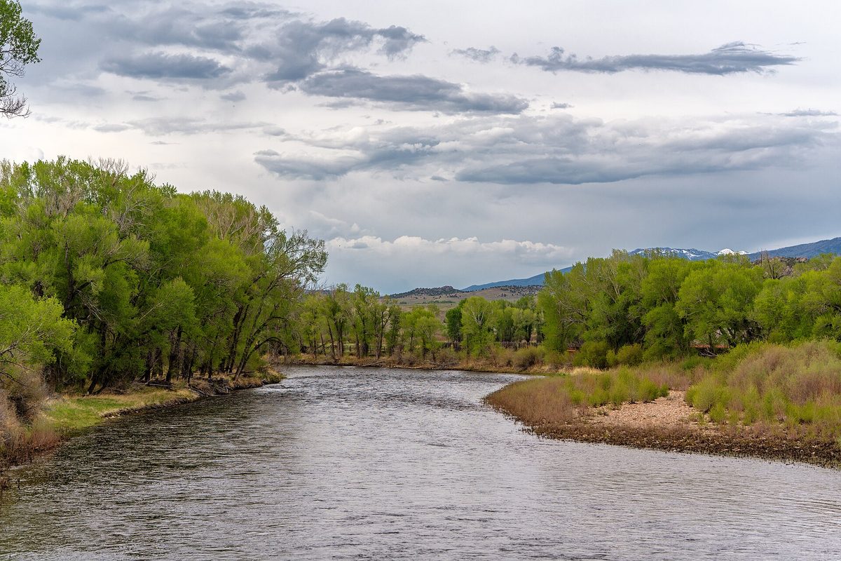 Wyoming river bending between cottonwood-lined banks and willow flats toward low hills and snow-streaked peaks under heavy gray clouds.