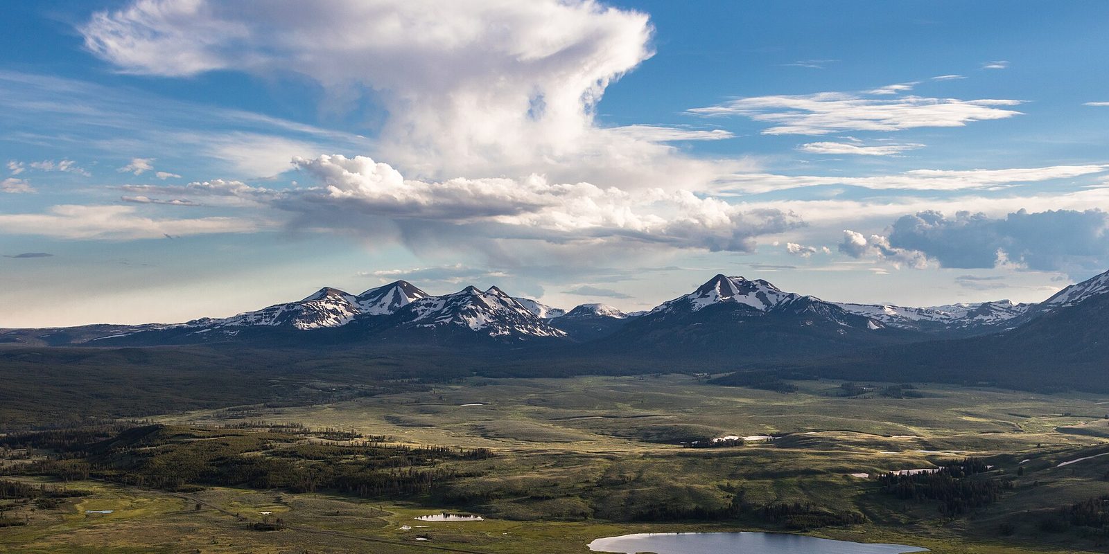 Snow-streaked peaks above a broad green valley, small lake below, tall clouds overhead